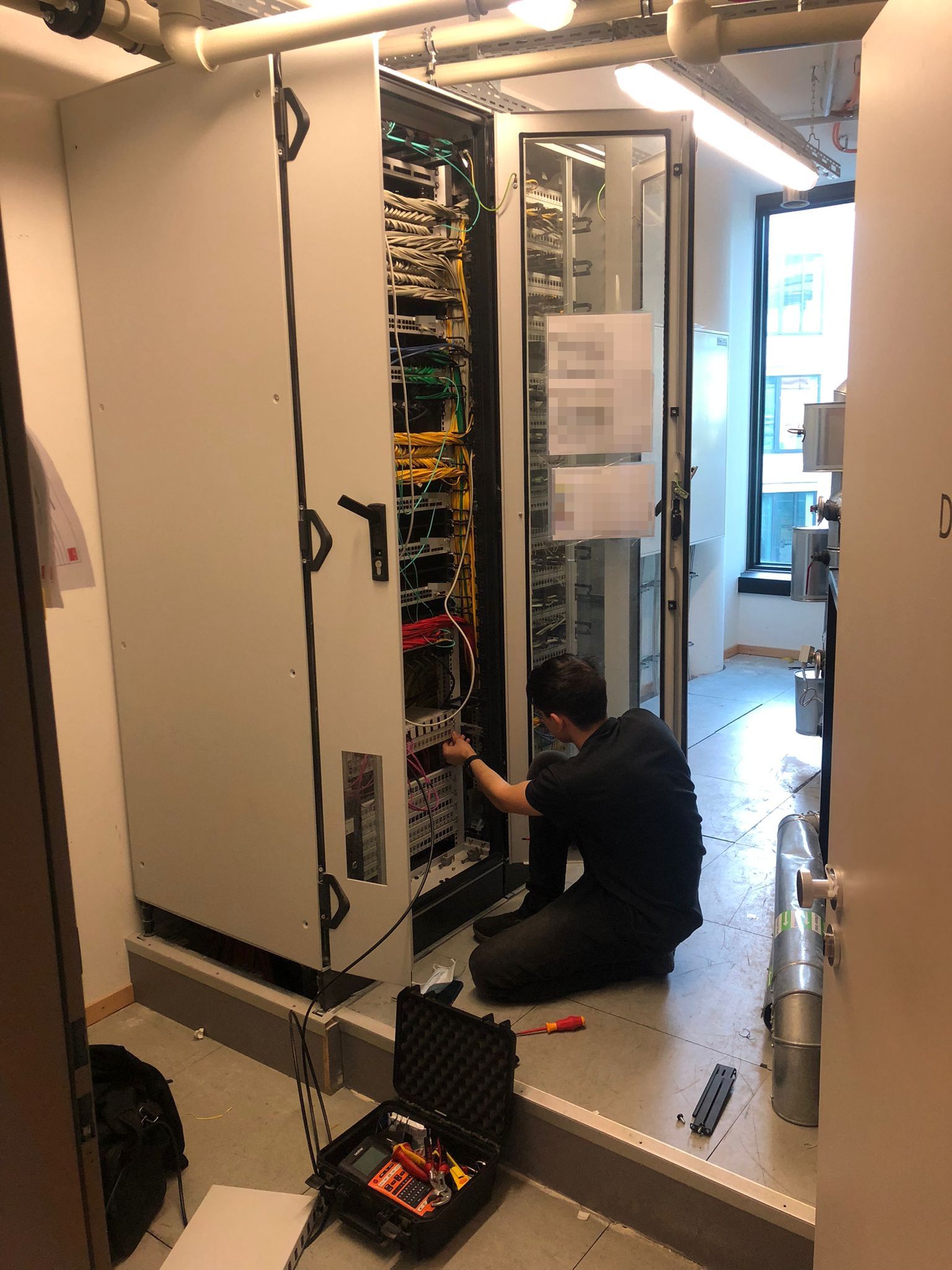 Techniker der TeKoNet kniet vor einem geöffneten Netzwerkschrank und arbeitet an einem Patchfeld, daneben ein geöffneter Gerätekoffer.