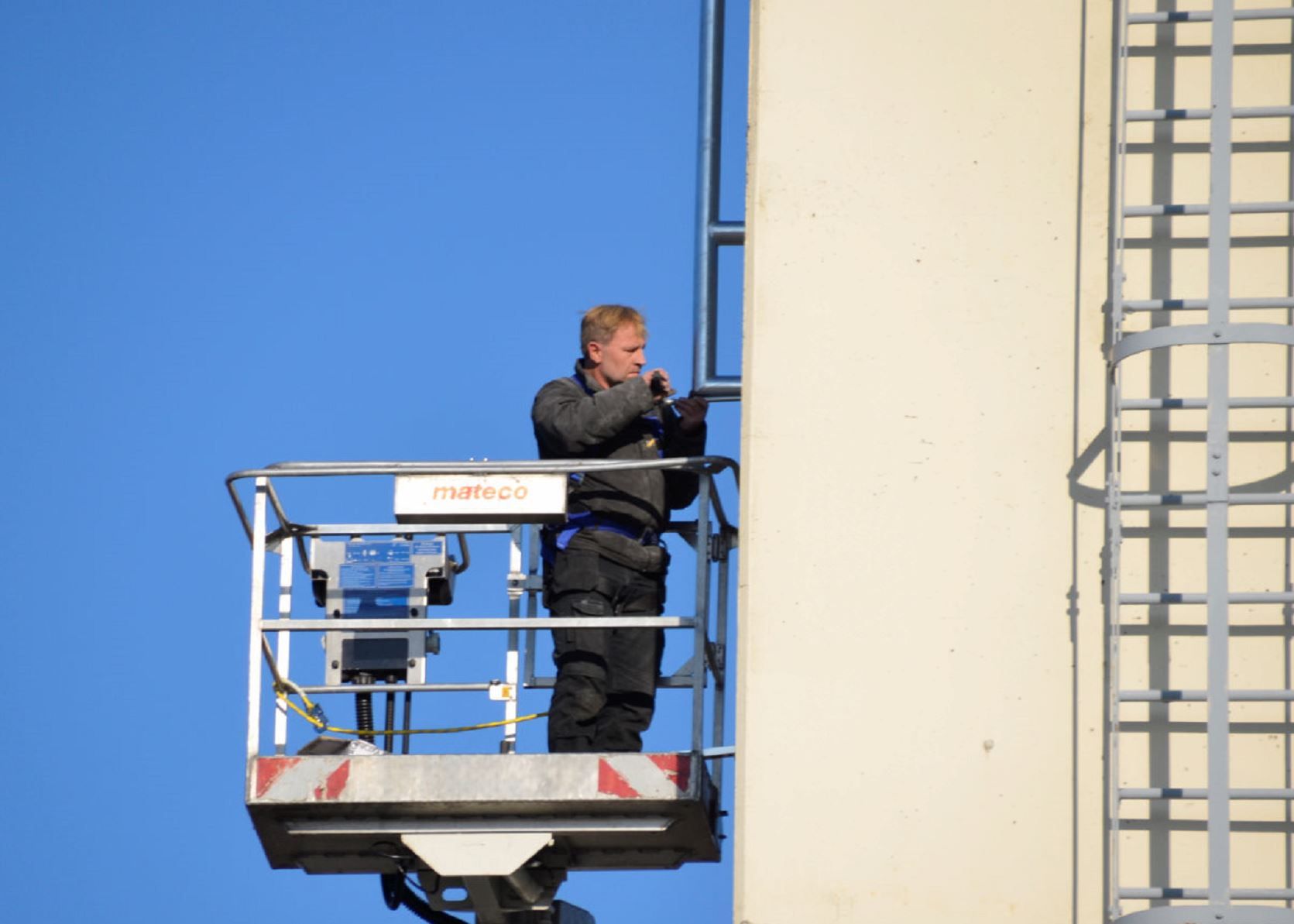 Techniker auf einer Hubarbeitsbühne montiert eine Richtfunkantenne an einer Gebäudewand vor blauem Himmel.