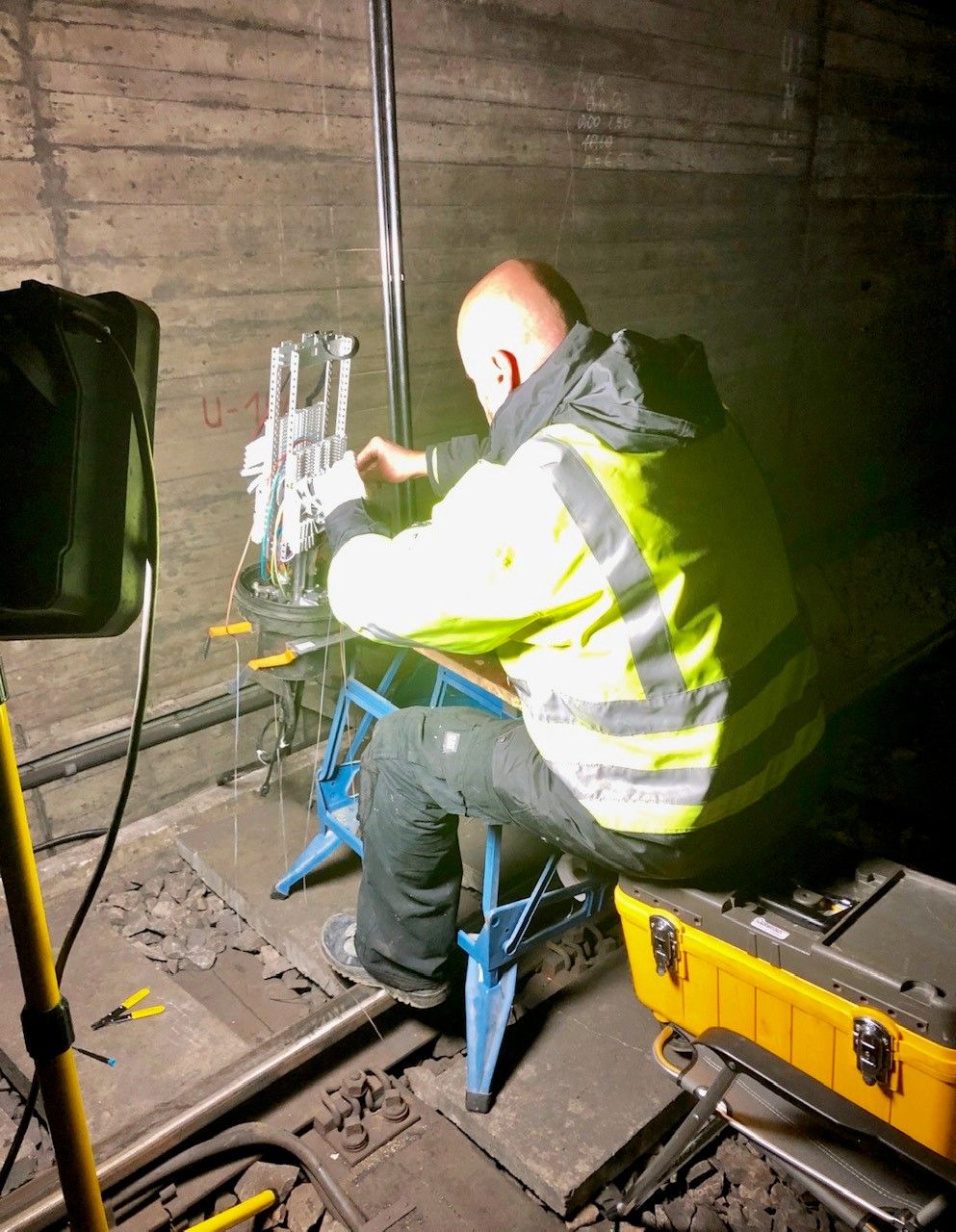 Techniker in Warnkleidung spleißt in einem Tunnel bei Baustellenbeleuchtung an einem Verteiler über den Gleisen.